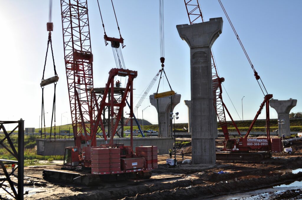 Construction site featuring two red Maxim Crane crawler cranes erecting large concrete bridge columns. The cranes are lifting and positioning concrete segments for the bridge structure. Workers are visible on site, guiding the placement of the segments.