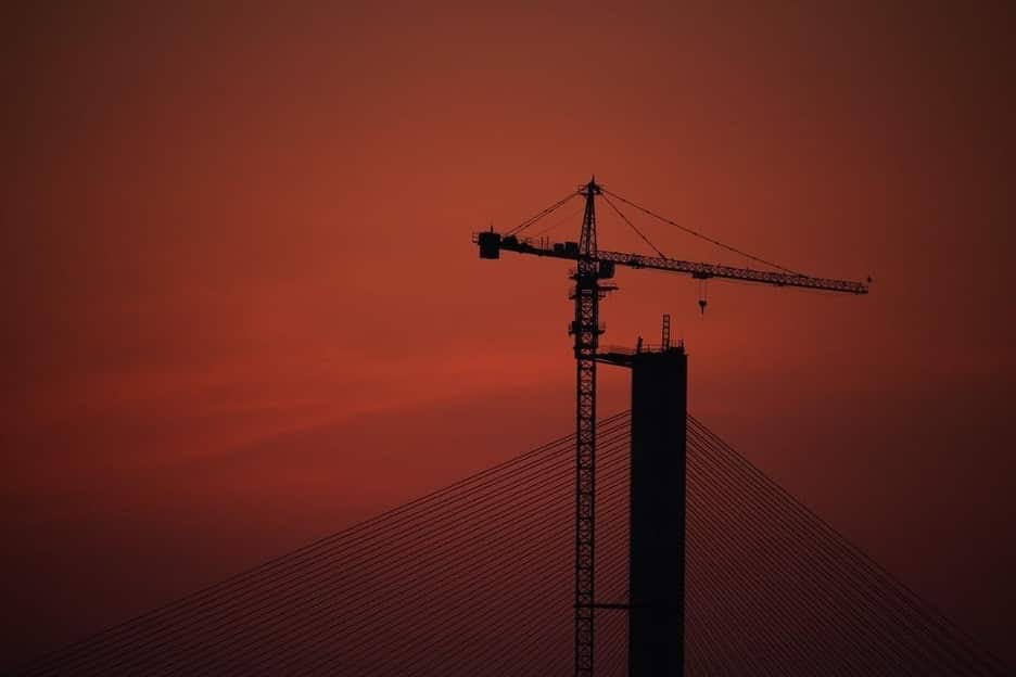 Silhouette of a tall construction crane against a vibrant red sunset sky, with cables from a bridge converging towards the base of the crane.