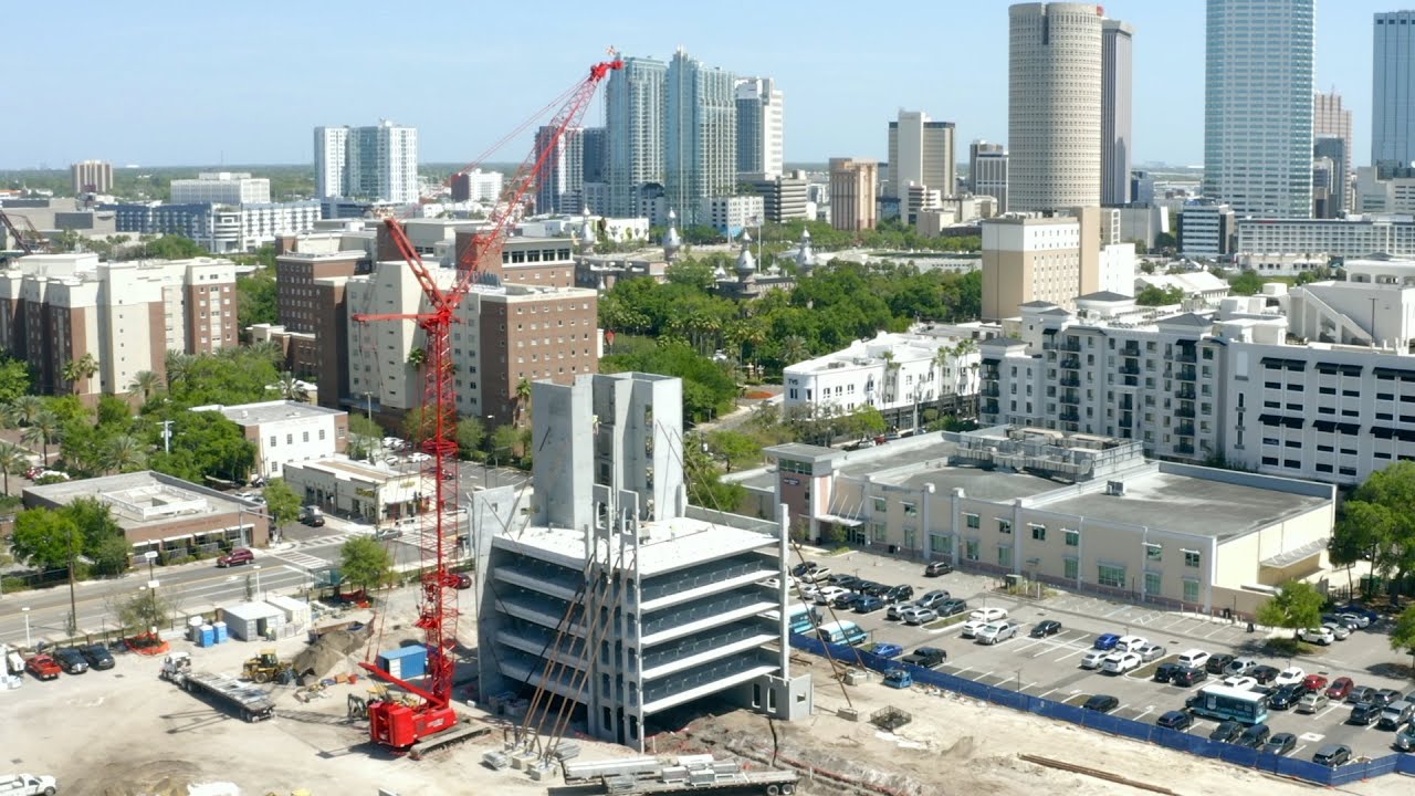 Aerial view of a construction site in an urban area with a prominent red Maxim Crane crawler crane in operation. The crane is being used to assemble a multi-story building. Surrounding the construction site are various residential and commercial buildings. In the background, the city skyline features tall skyscrapers and dense urban development.