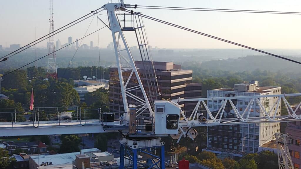 Close-up view of a large tower crane against a backdrop of a cityscape. The crane’s metal framework and control cab are prominently featured, with various cables and components attached. In the background, several multi-story buildings and lush green trees are visible, with the hazy city skyline in the distance under a clear sky.