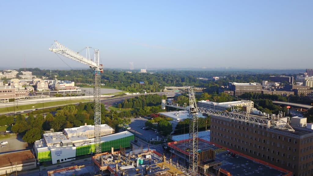 Aerial view of a construction site with prominent tower cranes in operation. The cranes tower over the skeletal framework of a building under construction in the foreground. The background contains a mix of commercial buildings, green spaces, and a highway, with a clear sky providing visibility of the distant tree-lined horizon.