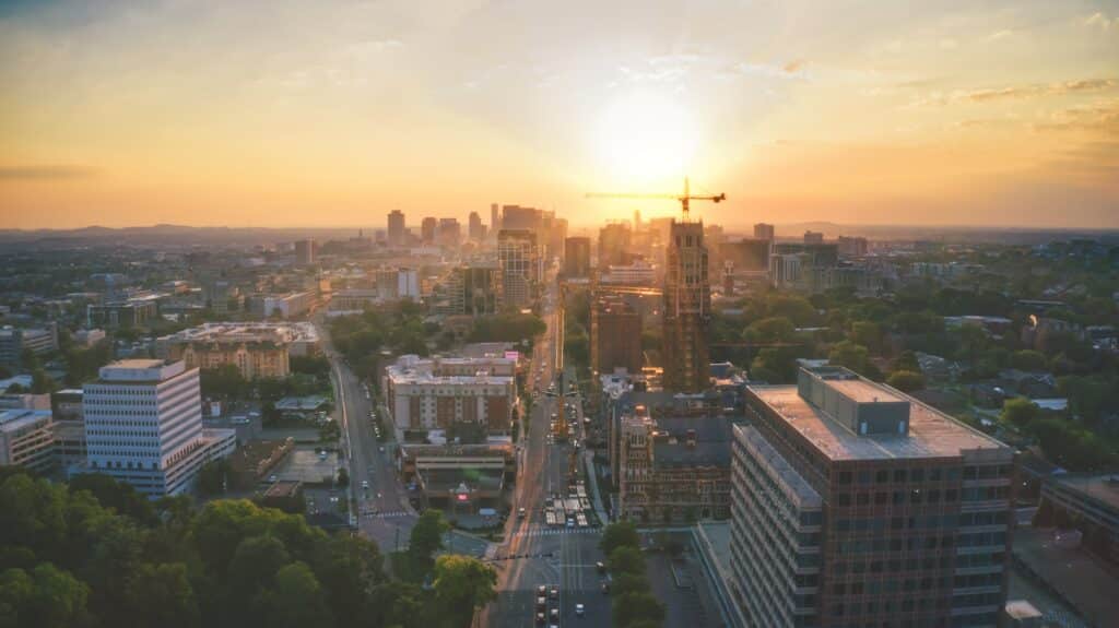 Aerial view of a city skyline at sunset, with the sun casting a warm glow over the buildings. A prominent tower crane is visible near the center, indicating ongoing construction. The streets below are lined with various buildings, including modern office structures and residential areas, with lush green trees interspersed throughout the cityscape.