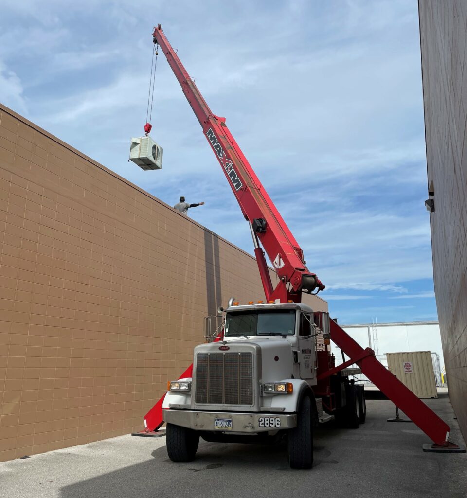 A Maxim Crane boom truck in a narrow alley, with its red boom extended vertically, lifting a large HVAC unit over a building. The truck is stabilized with extended outriggers. A person on the rooftop is directing the placement of the lifted unit. The truck cab is white with "Maxim Crane" branding on the side. The sky is partly cloudy with a mix of blue and white.