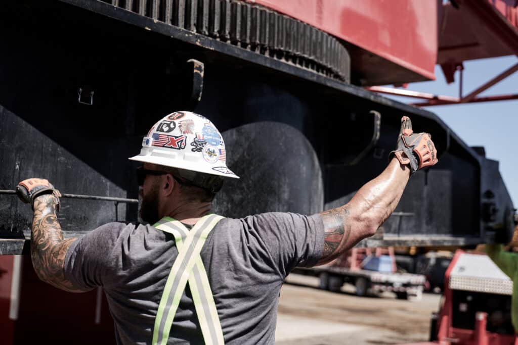 A Maxim Crane worker with a hard hat covered in stickers is adjusting equipment on a crane. He is wearing safety gloves and a gray shirt, with reflective straps visible. The background shows machinery and equipment on a jobsite, under clear blue skies.
