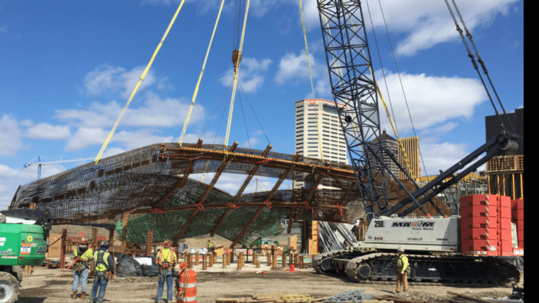 A construction jobsite featuring workers in safety gear overseeing the lifting of a large steel structure with cranes. The structure is partially lifted off the ground, surrounded by equipment and materials. In the background, there are tall buildings and cranes under a blue sky with scattered clouds.
