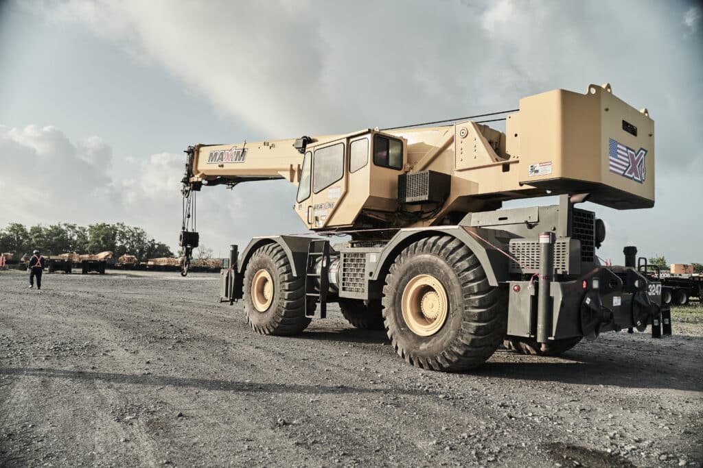 A yellow Maxim Crane rough terrain crane parked on a gravel lot under a cloudy sky. The crane features large, rugged tires suitable for uneven surfaces, and its boom is compacted. The company's logo is visible on both the boom and the rear section of the crane. Additional construction equipment and a person in a high-visibility vest can be seen in the background.