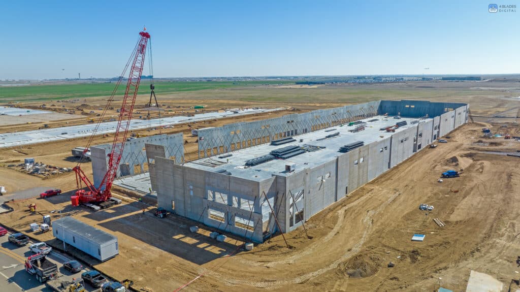 Aerial view of a construction site at Denali Logistics Park featuring a prominent red Maxim Crane crawler crane. The crane is lifting large precast concrete panels for the construction of a large industrial building. The building is in its early stages with walls being erected and construction materials spread out on the site.