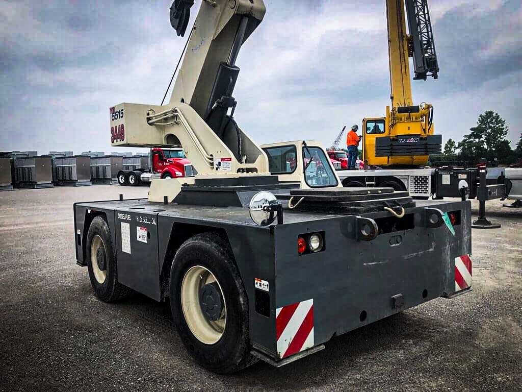 A Maxim Crane carry deck crane in a lot with other construction vehicles and equipment. The crane has a compact, maneuverable design with a flat deck and a telescoping boom. The backdrop includes a red truck and a yellow crane with workers, all under an overcast sky.