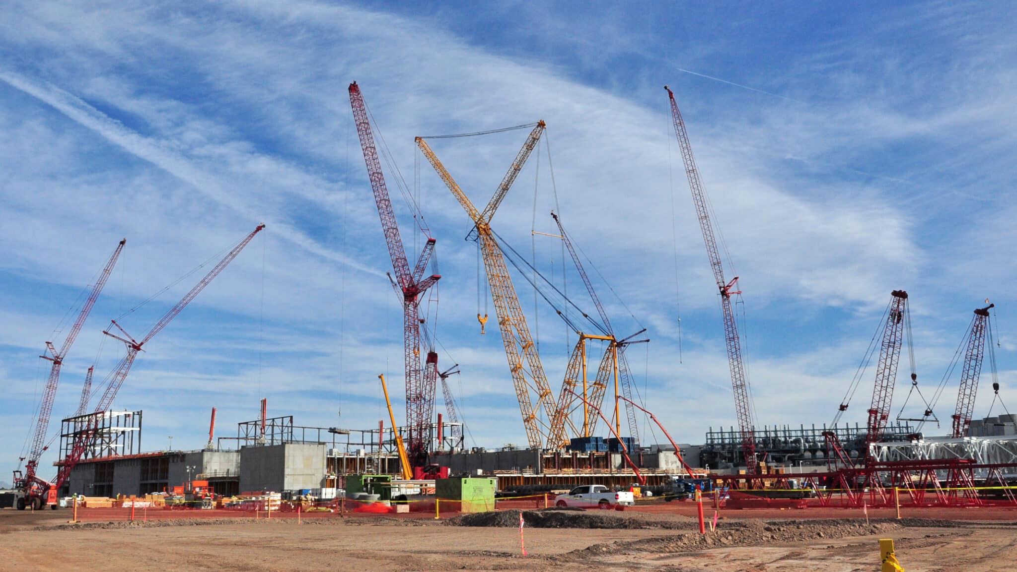 A bustling construction site at the Intel Fab 42 project, showcasing numerous towering red and yellow cranes from Maxim Crane Works. The cranes are actively engaged in lifting and positioning large steel beams and other construction materials for the building structures. Workers and various construction equipment are seen scattered throughout the expansive, dusty site. The sky is clear with streaks of clouds, indicating a bright and productive day.