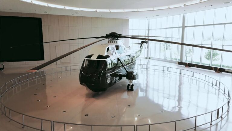 A large, Marine One helicopter is displayed inside a spacious, well-lit exhibition hall with floor-to-ceiling windows. The helicopter's body is predominantly dark in color with the white top and distinct markings associated with its presidential transport role. The helicopter is encircled by a safety railing, and the overall setting appears modern and pristine. This image is from a Maxim Crane lift project at the George Bush Presidential Library and Museum.