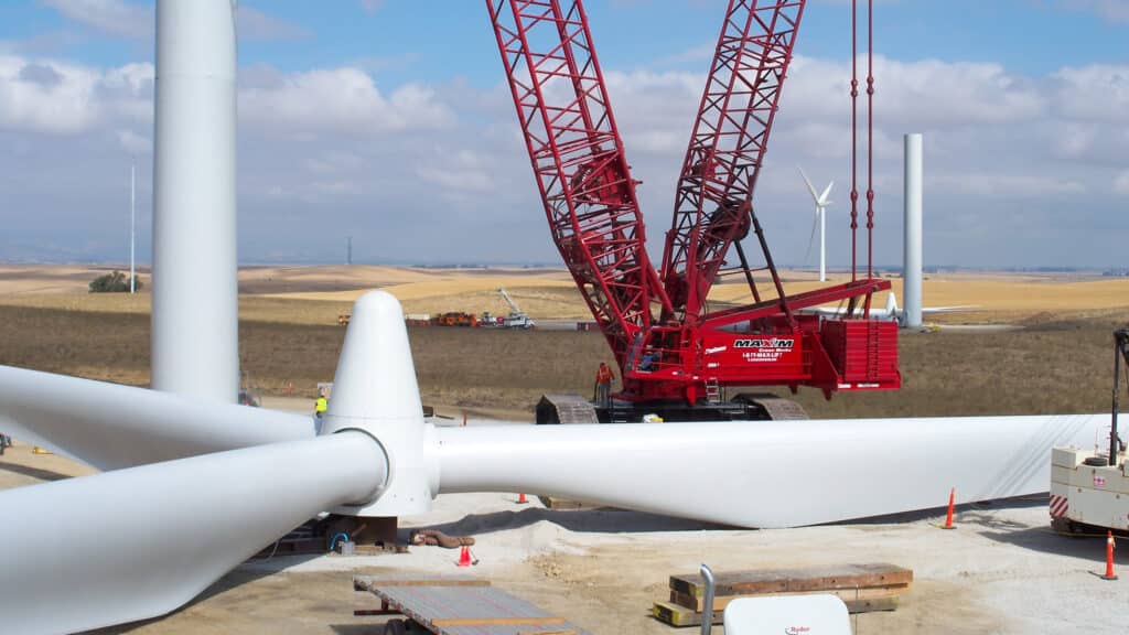 A Maxim Crane crawler crane is being used to assemble a wind turbine in an open field. The image shows the rotor blades of the turbine lying on the ground, ready to be lifted and attached to the tower. The landscape in the background features rolling hills and another wind turbine already standing. The sky is partly cloudy, and the area is surrounded by farmland. Construction equipment and workers are also visible in the vicinity.