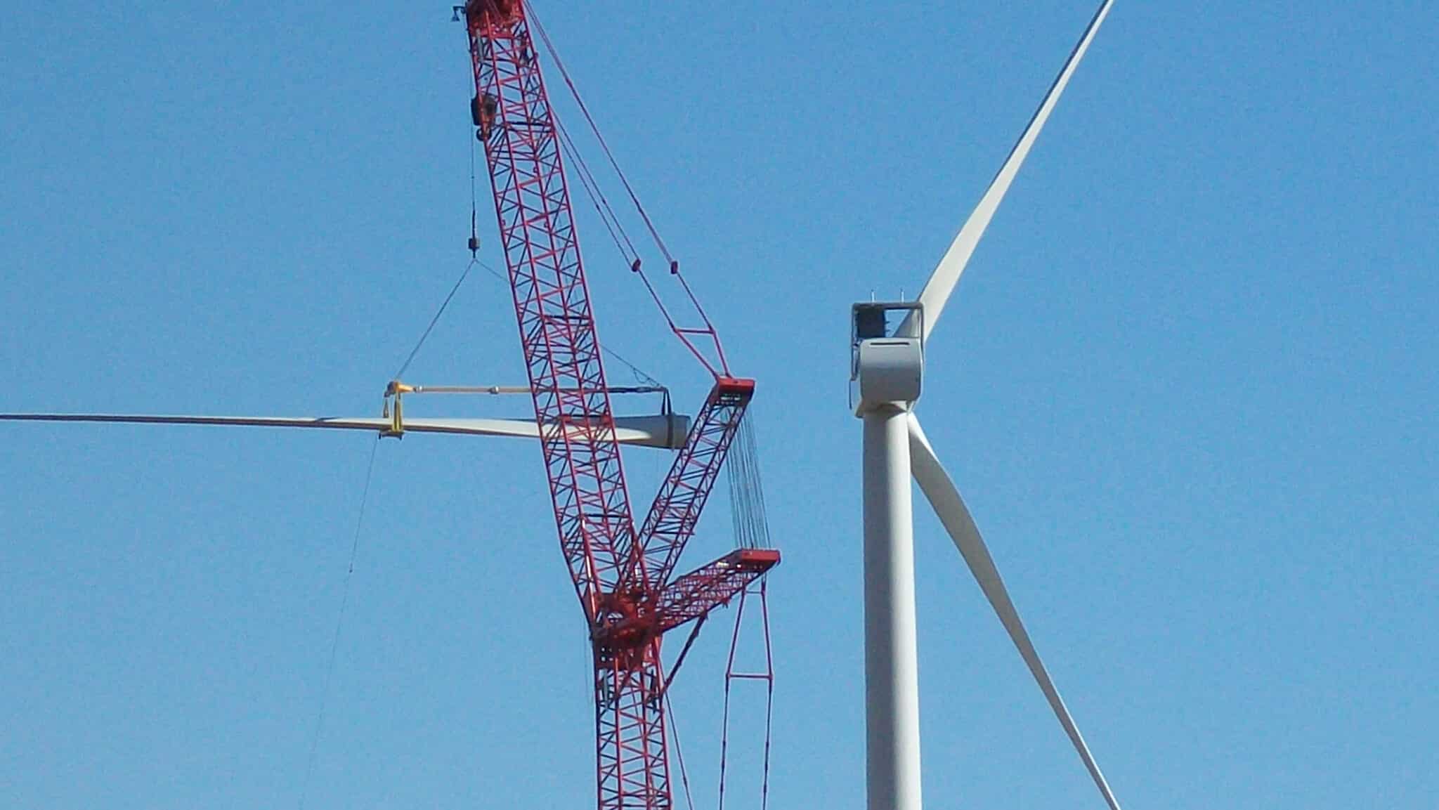 A red crane from Maxim Crane Works is carefully installing a blade onto a wind turbine at a wind farm. The crane's boom is extended high into the clear blue sky, precisely positioning the large, white turbine blade. The wind turbine's tall tower and other blades are prominently visible, highlighting the scale of the installation operation.