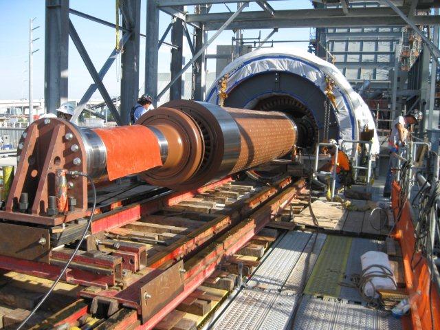The image shows an industrial setting where a heavy cylindrical component is being moved using the jack and slide method. The component is placed on a set of tracks with hydraulic jacks used to control and maneuver the slide. Several workers and scaffolding can be seen in the background, indicating an ongoing engineering or maintenance operation.