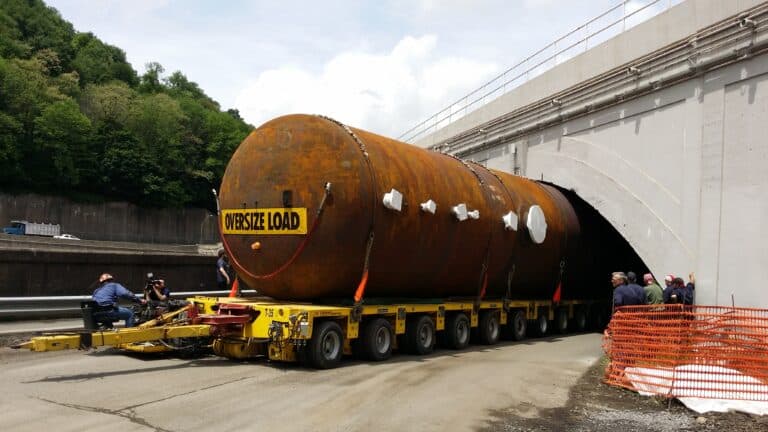 A Maxim Crane heavy haul vehicle transporting an oversized load through a tunnel, with workers observing the process. The load is secured with chains and marked as "OVERSIZE LOAD," emphasizing the complexity of moving large items safely.
